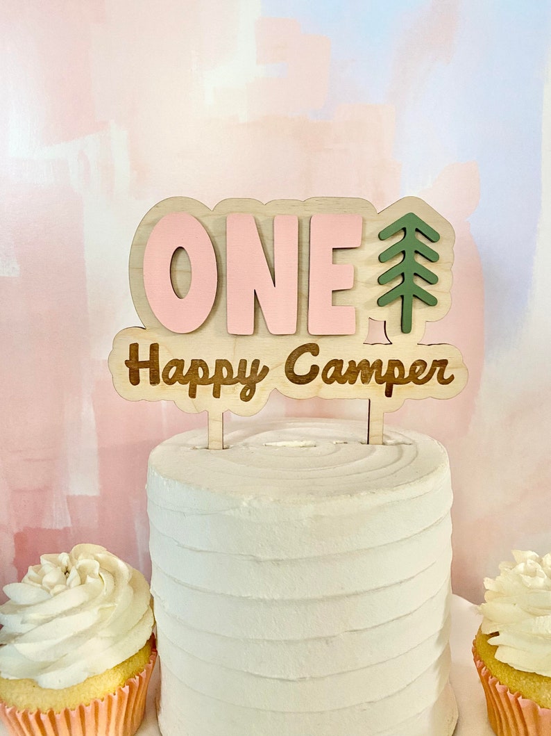 K&ouml;nnte beinhalten: Ein Holz-Kuchentopper mit dem Wort "ONE" in rosa Buchstaben und den Worten "Happy Camper" in braunen Buchstaben. Rechts neben dem Wort "ONE" befindet sich eine gr&uuml;ne Baumsilhouette.