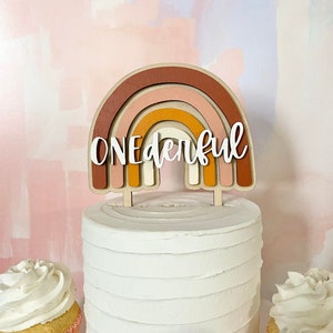 May include: A white cake with a wooden rainbow topper that says "ONEderful". The rainbow is made of layered wood in shades of brown, orange, pink, and white.