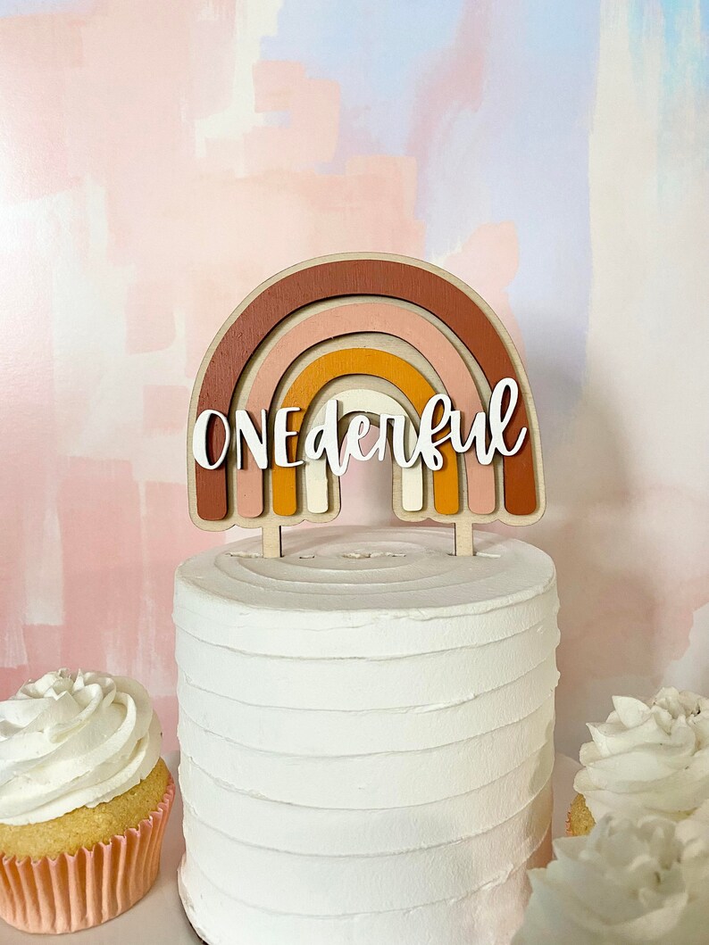 May include: A white cake with a wooden rainbow topper that says "ONEderful". The rainbow is made of layered wood in shades of brown, orange, pink, and white.