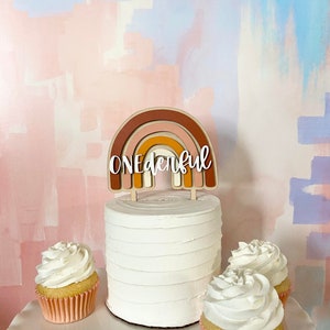 May include: A white cake with a rainbow-colored wooden topper that says "ONEderful". The cake is decorated with white frosting and is surrounded by three cupcakes with white frosting.