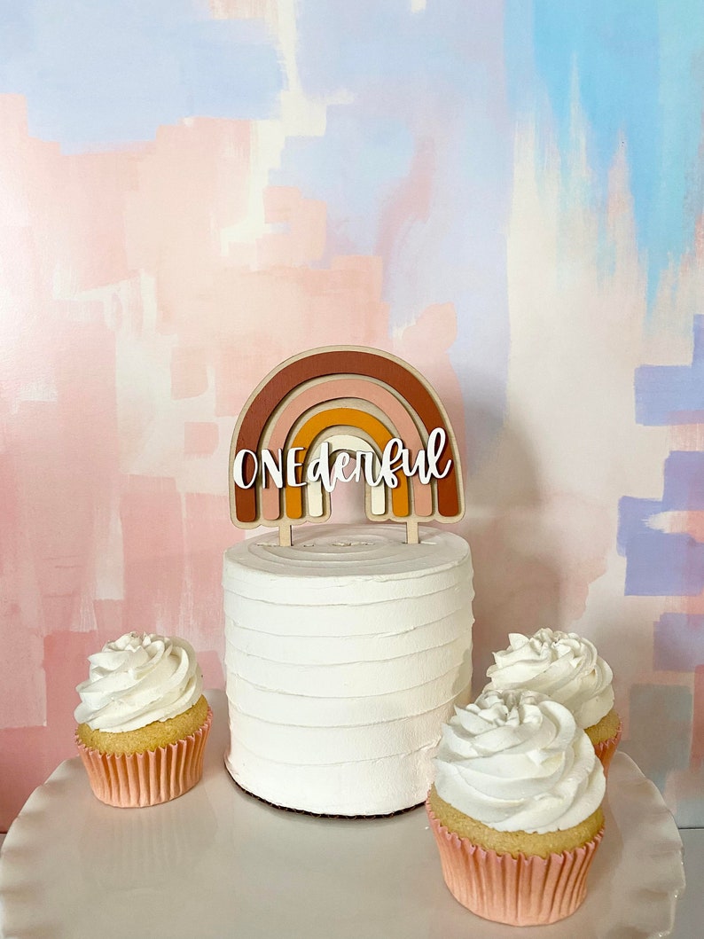 May include: A white cake with a rainbow-colored wooden topper that says "ONEderful". The cake is decorated with white frosting and is surrounded by three cupcakes with white frosting.