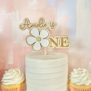 Pu&ograve; includere: Una torta bianca glassata con un cake topper in legno che recita "Amelie is ONE". Il cake topper presenta un design a margherita. Due cupcakes con glassa bianca sono ai lati della torta, in pirottini rosa.