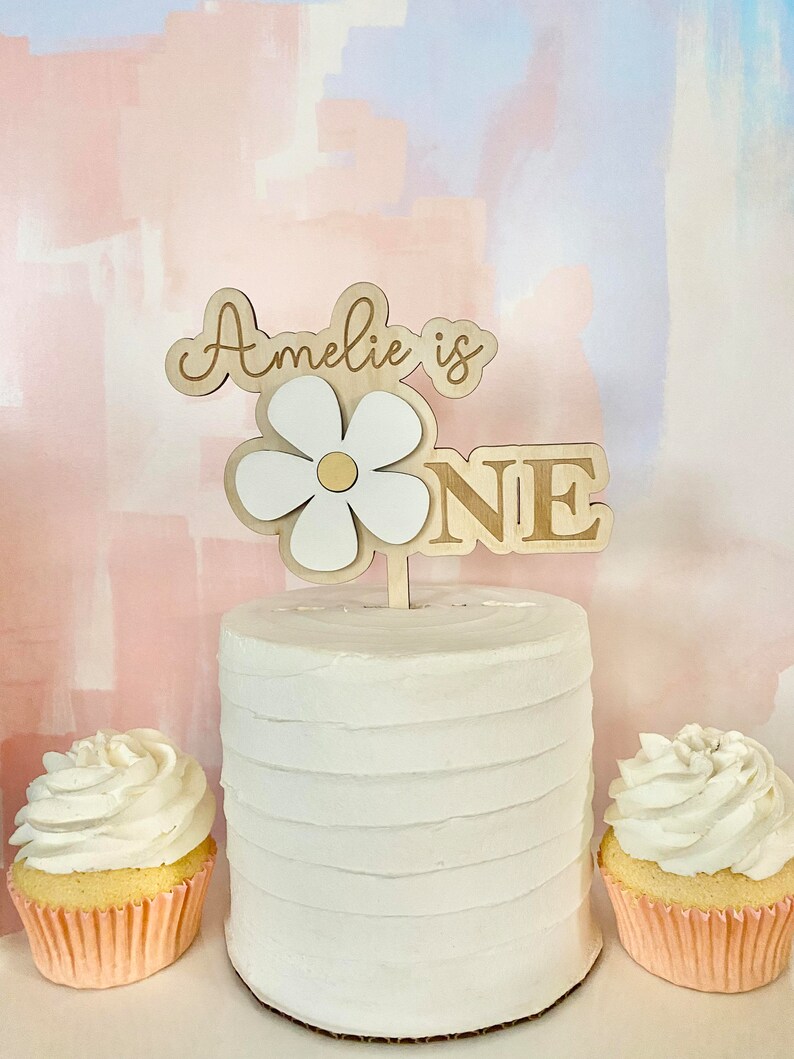 Pu&ograve; includere: Una torta bianca glassata con un cake topper in legno che recita "Amelie is ONE". Il cake topper presenta un design a margherita. Due cupcakes con glassa bianca sono ai lati della torta, in pirottini rosa.