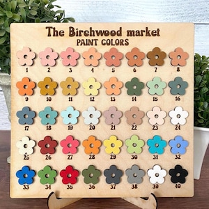 May include: A wooden display board with the text "The Birchwood market PAINT COLORS" features 40 flower-shaped paint color samples. The colors range from light pink to dark brown, with various shades of blue, green, and orange.
