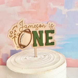 May include: A wooden cake topper with the words "Jameson is ONE" in a mix of script and block letters. The "O" is replaced with a football graphic. The topper is on a white frosted cake, with a colorful background.