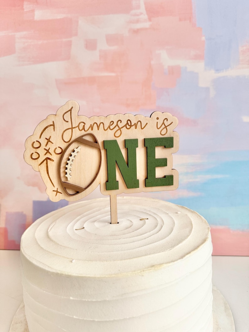 May include: A wooden cake topper with the words "Jameson is ONE" in a mix of script and block letters. The "O" is replaced with a football graphic. The topper is on a white frosted cake, with a colorful background.