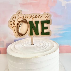 May include: A wooden cake topper with the text "Jameson is ONE" in a football theme. The "ONE" is in green block letters. The topper is placed on a white frosted cake.