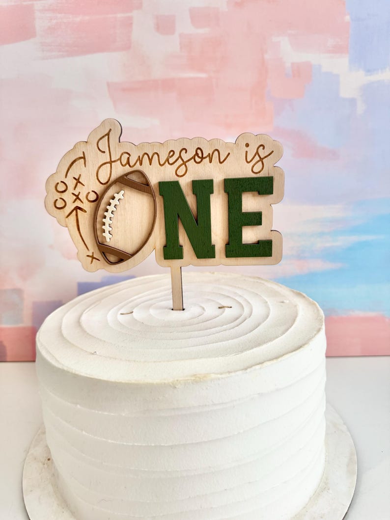 May include: A wooden cake topper with the text "Jameson is ONE" in a football theme. The "ONE" is in green block letters. The topper is placed on a white frosted cake.