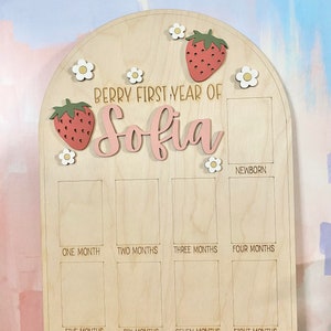 Puede incluir: Tablero de hitos de madera con tema de fresas y margaritas. El tablero est&aacute; grabado con el texto "Berry First Year of Sofia" y tiene espacios para fotos mensuales. Incluye un espacio para fotos de reci&eacute;n nacidos.