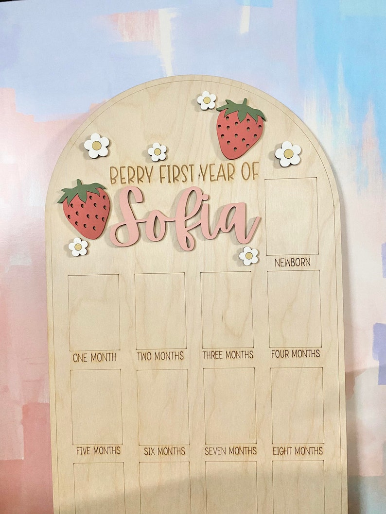 Puede incluir: Tablero de hitos de madera con tema de fresas y margaritas. El tablero est&aacute; grabado con el texto "Berry First Year of Sofia" y tiene espacios para fotos mensuales. Incluye un espacio para fotos de reci&eacute;n nacidos.
