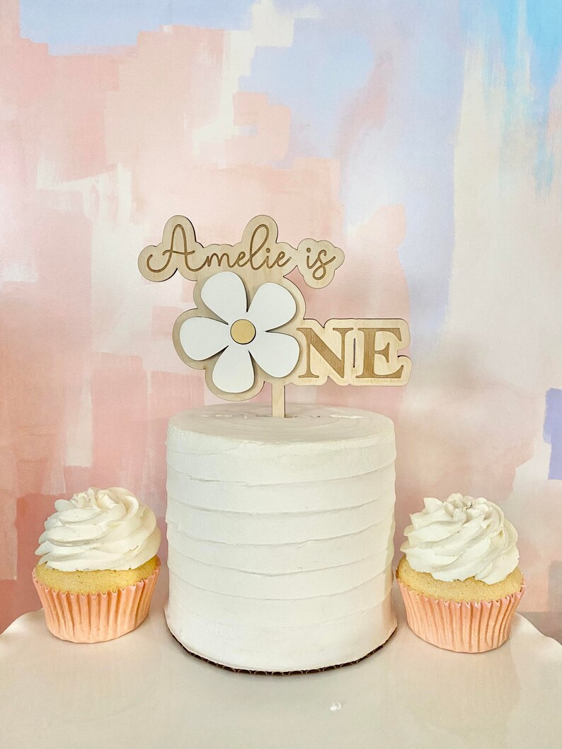 Pu&ograve; includere: Una torta bianca glassata con un cake topper a forma di margherita in legno che recita "Amelie is ONE". Due cupcakes con glassa bianca si trovano su entrambi i lati della torta. Lo sfondo &egrave; un design astratto rosa e blu pastello.