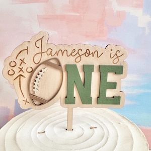 May include: A wooden cake topper with the words "Jameson is ONE". The topper features a football and play diagram on the left and green letters spelling "ONE" on the right. The topper is placed on a white frosted cake.