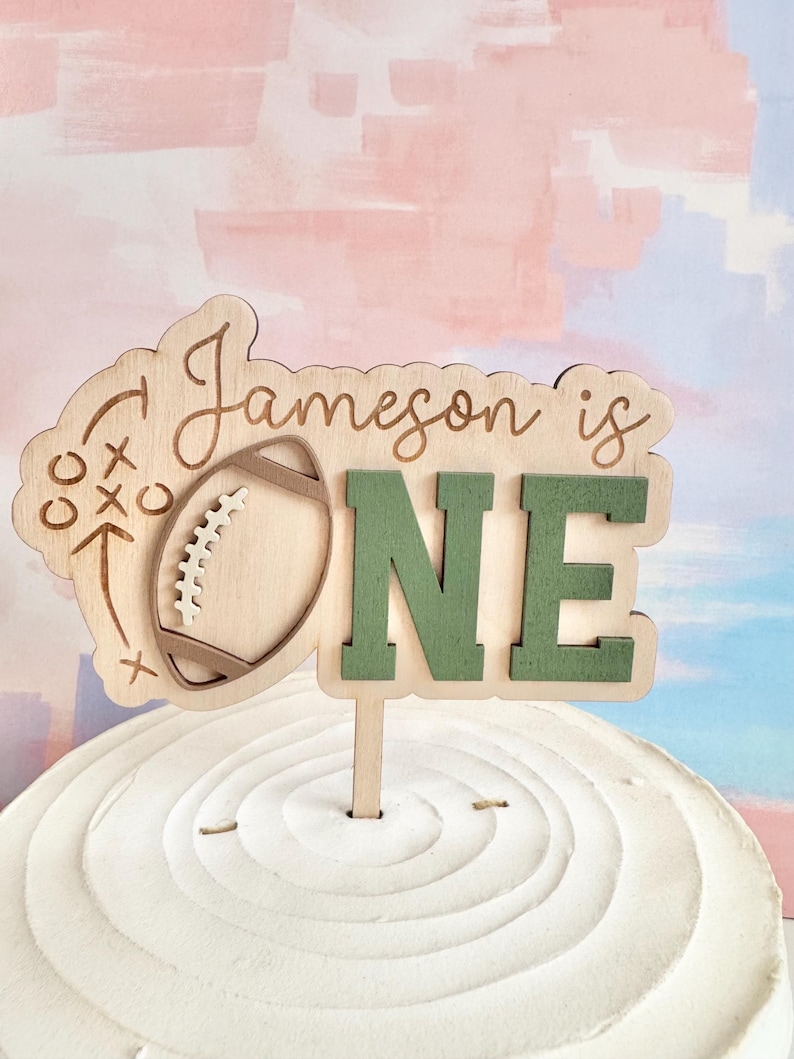 May include: A wooden cake topper with the words "Jameson is ONE". The topper features a football and play diagram on the left and green letters spelling "ONE" on the right. The topper is placed on a white frosted cake.