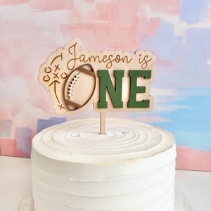 May include: A wooden cake topper with the text "Jameson is ONE" in a playful font. The design includes a football and play diagrams. The topper is placed on a white frosted cake, perfect for a first birthday celebration.