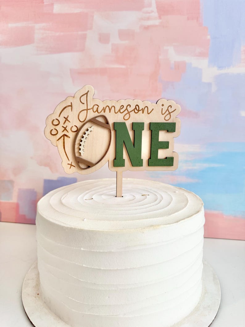 May include: A wooden cake topper with the text "Jameson is ONE" in a playful font. The design includes a football and play diagrams. The topper is placed on a white frosted cake, perfect for a first birthday celebration.