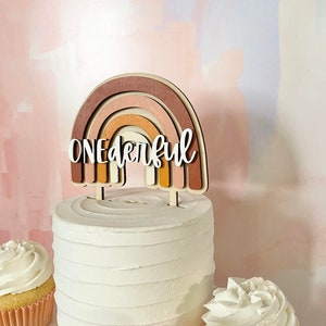 May include: A white cake with a wooden rainbow topper that says "One-derful". The cake is decorated with white frosting and is surrounded by two cupcakes with white frosting.