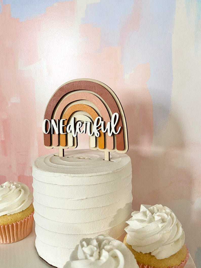 May include: A white cake with a wooden rainbow topper that says "One-derful". The cake is decorated with white frosting and is surrounded by two cupcakes with white frosting.