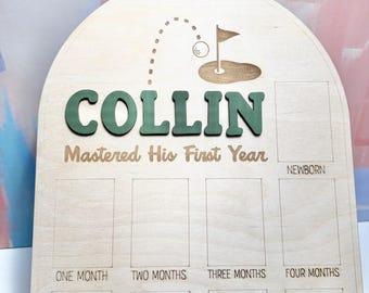 May include: Wooden milestone board with the name "COLLIN" in green letters. The board features a golf-themed design and spaces to document a baby's first year, including "Newborn" and "One Month" to "Eight Months".