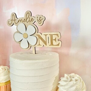 Pu&ograve; includere: Un cake topper in legno con le parole "Amelie is ONE" e un design a margherita. Il cake topper &egrave; posto su una torta glassata bianca. Due cupcakes con glassa bianca sono su entrambi i lati della torta.