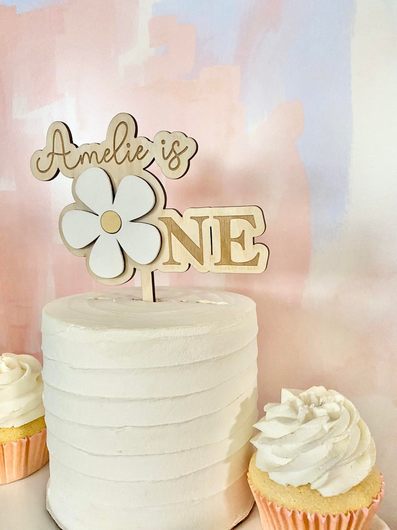 Pu&ograve; includere: Un cake topper in legno con le parole "Amelie is ONE" e un design a margherita. Il cake topper &egrave; posto su una torta glassata bianca. Due cupcakes con glassa bianca sono su entrambi i lati della torta.
