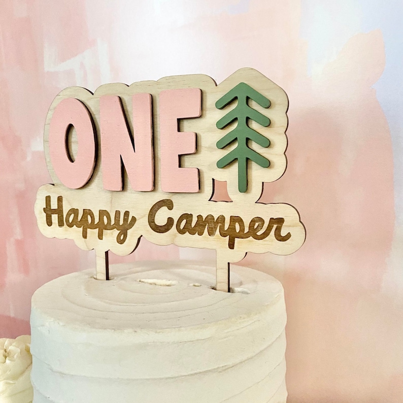 K&ouml;nnte beinhalten: Ein rosa und brauner Holz-Kuchentopper mit dem Wort "ONE" und dem Text "Happy Camper" mit einem gr&uuml;nen Tannenbaum-Design.