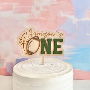 May include: A wooden cake topper with the text "Jameson is ONE." The topper features a football design and is set on a white frosted cake. The background has pink, blue, and white brushstrokes.