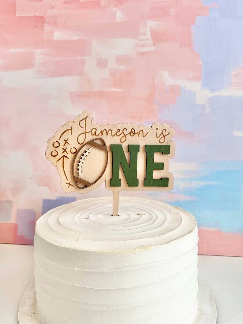 May include: A wooden cake topper with the text "Jameson is ONE." The topper features a football design and is set on a white frosted cake. The background has pink, blue, and white brushstrokes.