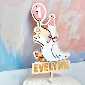 May include: A wooden cake topper featuring a cartoon goose holding a pink balloon with the number 1. The goose wears a party hat and a pink bow. The name "Evelynn" is displayed on a wooden sign below the goose.