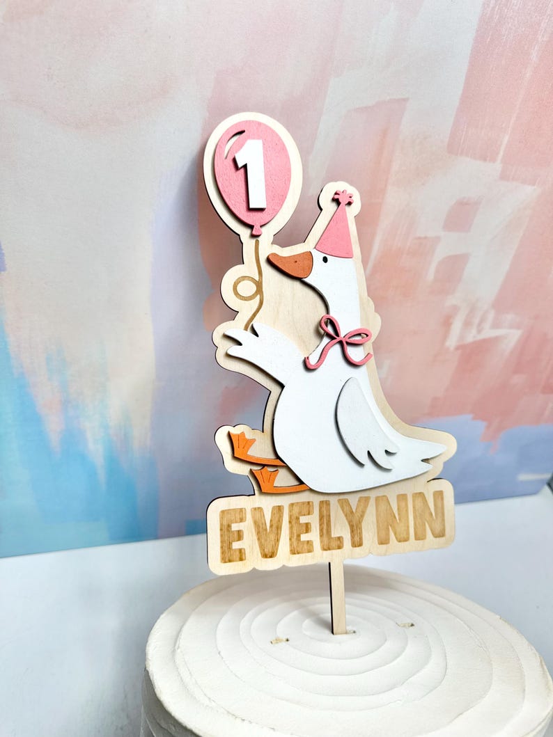 May include: A wooden cake topper featuring a cartoon goose holding a pink balloon with the number 1. The goose wears a party hat and a pink bow. The name "Evelynn" is displayed on a wooden sign below the goose.