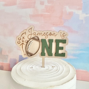 May include: A wooden cake topper with the text "Jameson is ONE" and a football design. The letters "ONE" are green. The topper is placed on a white frosted cake with a textured surface. The background is a pastel color wash.