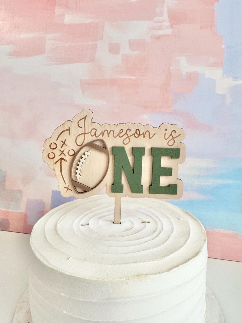 May include: A wooden cake topper with the text "Jameson is ONE" and a football design. The letters "ONE" are green. The topper is placed on a white frosted cake with a textured surface. The background is a pastel color wash.