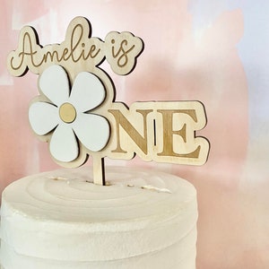 Pu&ograve; includere: Un cake topper in legno con la scritta "Amelie is ONE". Il topper presenta una margherita con petali bianchi e un centro dorato. La torta &egrave; bianca con glassa strutturata.