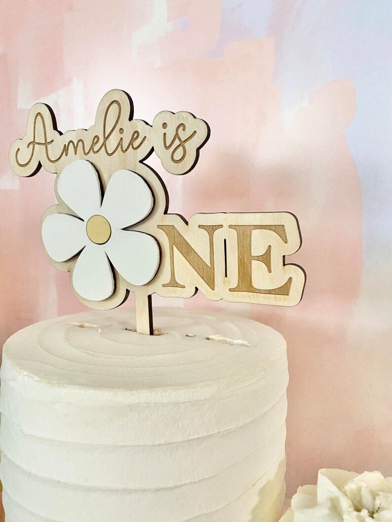 Pu&ograve; includere: Un cake topper in legno con la scritta "Amelie is ONE". Il topper presenta una margherita con petali bianchi e un centro dorato. La torta &egrave; bianca con glassa strutturata.