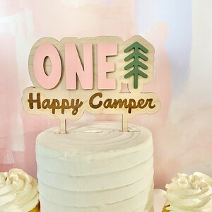 K&ouml;nnte beinhalten: Ein rosafarbener und brauner Holz-Kuchentopper mit dem Wort "ONE" und einem gr&uuml;nen Tannenbaum. Der Text "Happy Camper" steht unter der Zahl eins.