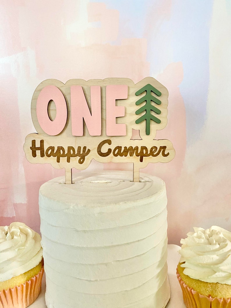 K&ouml;nnte beinhalten: Ein rosafarbener und brauner Holz-Kuchentopper mit dem Wort "ONE" und einem gr&uuml;nen Tannenbaum. Der Text "Happy Camper" steht unter der Zahl eins.