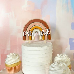 May include: A white cake with a rainbow-shaped cake topper that says "ONEderful". The cake is surrounded by three cupcakes with white frosting.