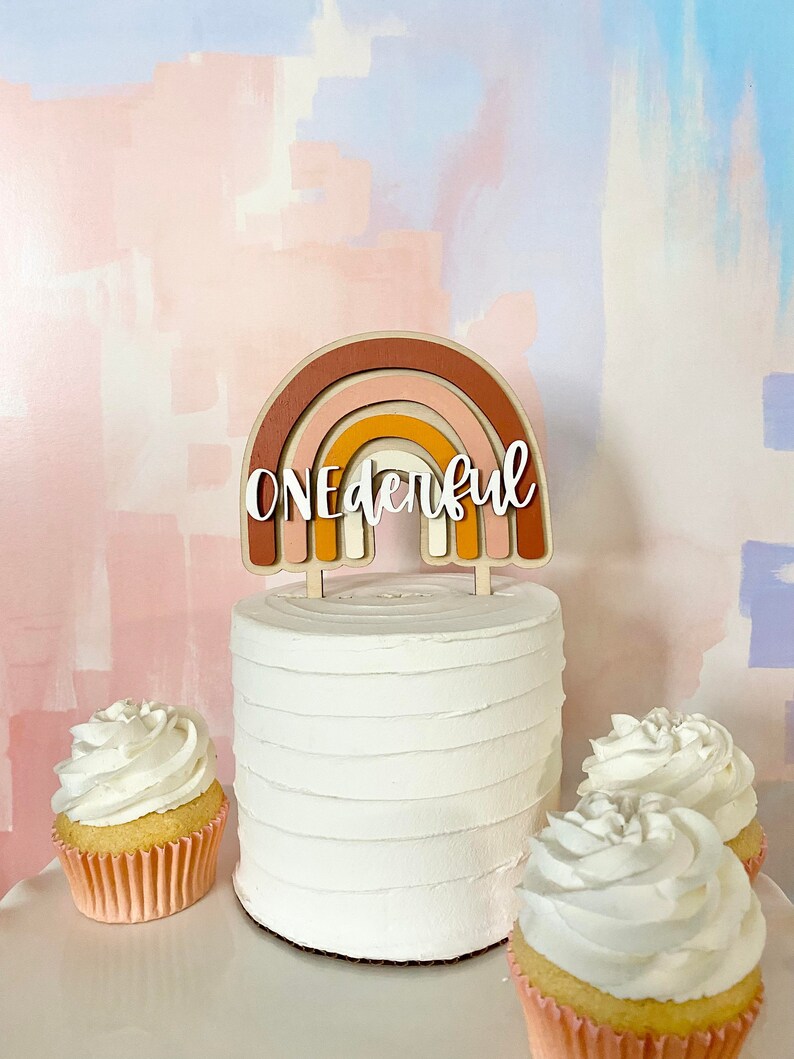 May include: A white cake with a rainbow-shaped cake topper that says "ONEderful". The cake is surrounded by three cupcakes with white frosting.