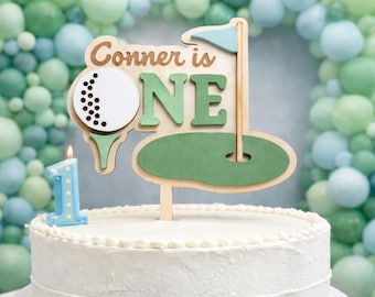 May include: A white frosted cake with a golf-themed cake topper that reads "Conner is ONE." The topper features a golf ball, tee, flag, and green. A blue number one candle is lit on the cake. The background has a balloon arch in shades of green and blue.