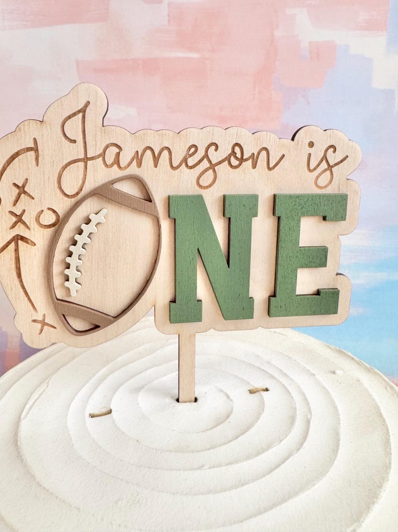 May include: A wooden cake topper with the text "Jameson is ONE." The topper features a football and play diagram design. The word "ONE" is in green, and the other elements are in natural wood tones. The topper is inserted into a white frosted cake.