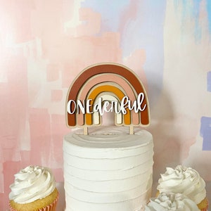 May include: A white cake with a wooden rainbow cake topper that says "ONEderful". The cake is decorated with white frosting and is surrounded by two cupcakes with white frosting.
