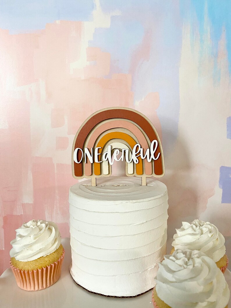 May include: A white cake with a wooden rainbow cake topper that says "ONEderful". The cake is decorated with white frosting and is surrounded by two cupcakes with white frosting.