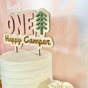 K&ouml;nnte beinhalten: Ein rosa-gr&uuml;ner Holz-Kuchentopper mit dem Wort "ONE" und einem Kiefernbaum-Design. Der Topper sagt auch "Happy Camper".