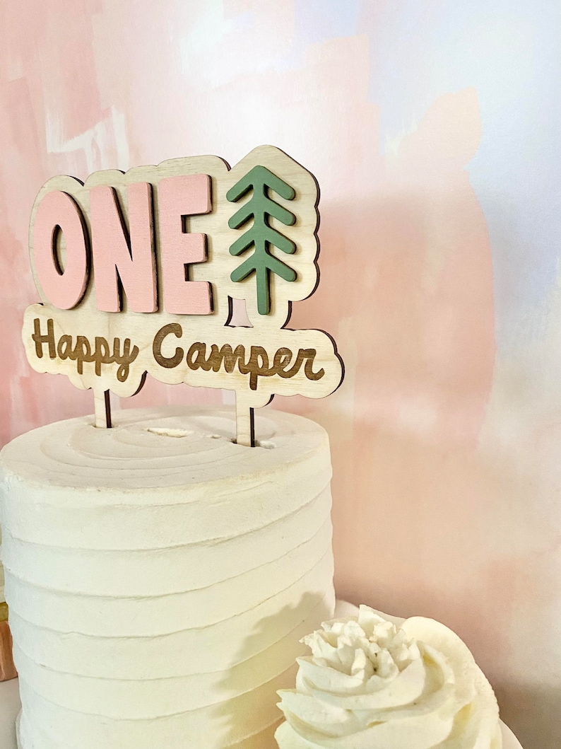 K&ouml;nnte beinhalten: Ein rosa-gr&uuml;ner Holz-Kuchentopper mit dem Wort "ONE" und einem Kiefernbaum-Design. Der Topper sagt auch "Happy Camper".