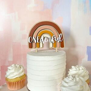 May include: A white cake with a wooden rainbow topper that says "ONEderful". The cake is decorated with white frosting and is surrounded by three cupcakes with white frosting.