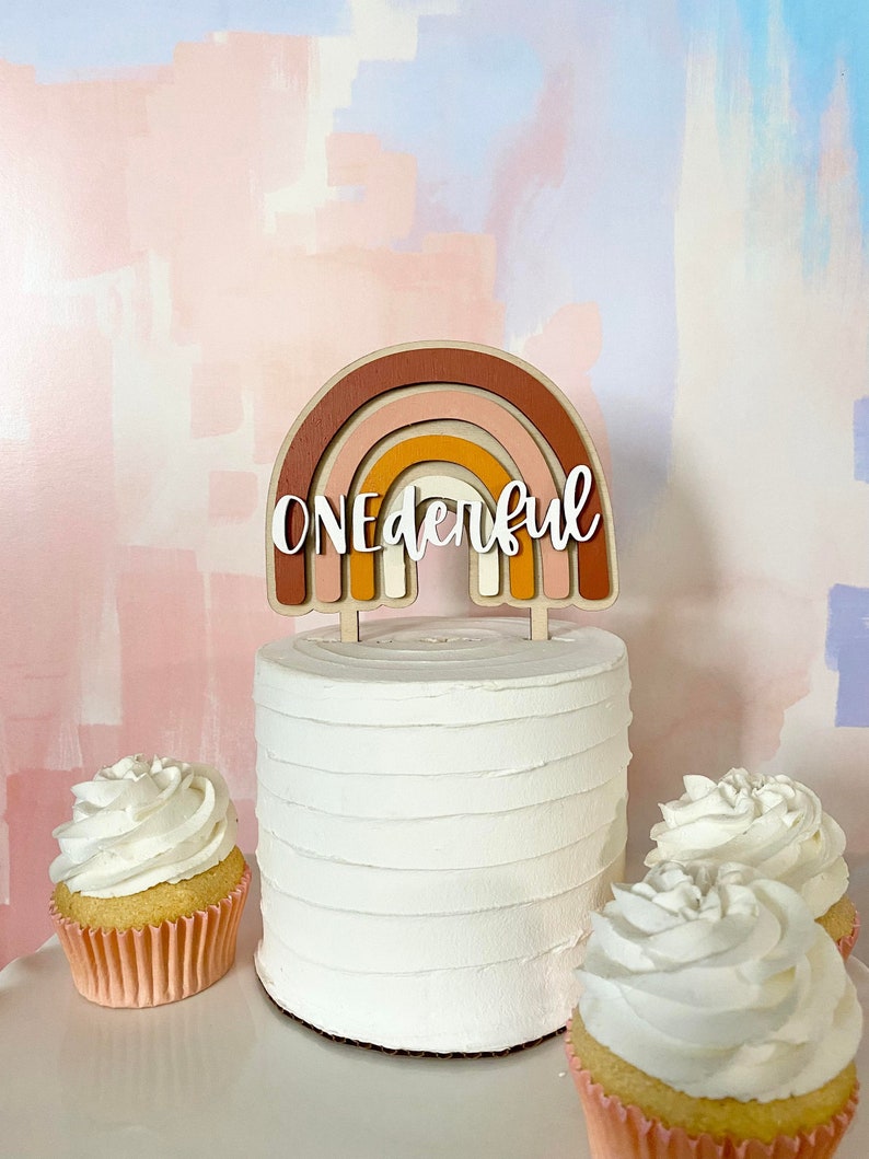 May include: A white cake with a wooden rainbow topper that says "ONEderful". The cake is decorated with white frosting and is surrounded by three cupcakes with white frosting.