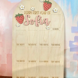 Puede incluir: Tablero de hitos de madera con el texto "Berry First Year of Sofia". Decorado con motivos de fresas y flores. Incluye espacios para fotos mensuales desde reci&eacute;n nacido hasta un a&ntilde;o.
