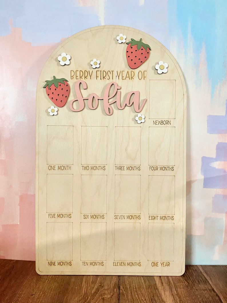 Puede incluir: Tablero de hitos de madera con el texto "Berry First Year of Sofia". Decorado con motivos de fresas y flores. Incluye espacios para fotos mensuales desde reci&eacute;n nacido hasta un a&ntilde;o.