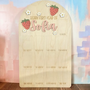 Puede incluir: Tablero de hitos de madera con tema de fresa. El tablero tiene forma de arco en la parte superior y presenta el texto "Berry First Year of Sofia". Incluye espacios para fotos mensuales y est&aacute; decorado con fresas y flores.
