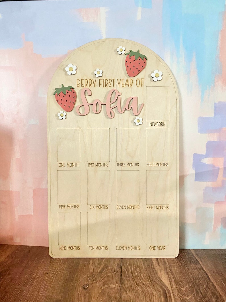 Puede incluir: Tablero de hitos de madera con tema de fresa. El tablero tiene forma de arco en la parte superior y presenta el texto "Berry First Year of Sofia". Incluye espacios para fotos mensuales y est&aacute; decorado con fresas y flores.