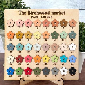 May include: A wooden display board with the text "The Birchwood market PAINT COLORS" features 40 flower-shaped paint color samples. The colors range from light pink to dark brown, with additional shades of orange, green, blue, and gray.